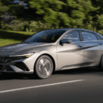 2025 Hyundai Elantra sedan in silver driving on a tree-lined road in daylight.