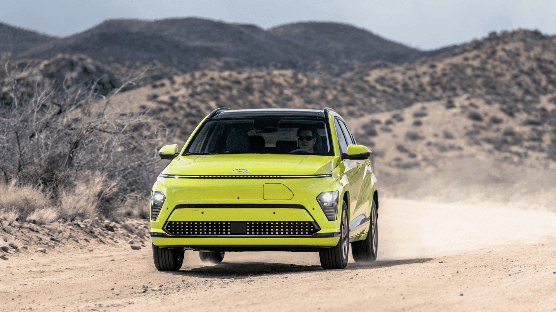 Lime green 2025 Hyundai Kona Electric SUV driving through a desert terrain head-on view
