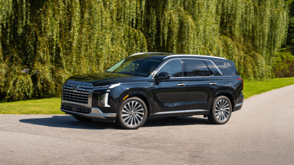2025 Hyundai Palisade in black driving on a sunny road with lush green trees in the background