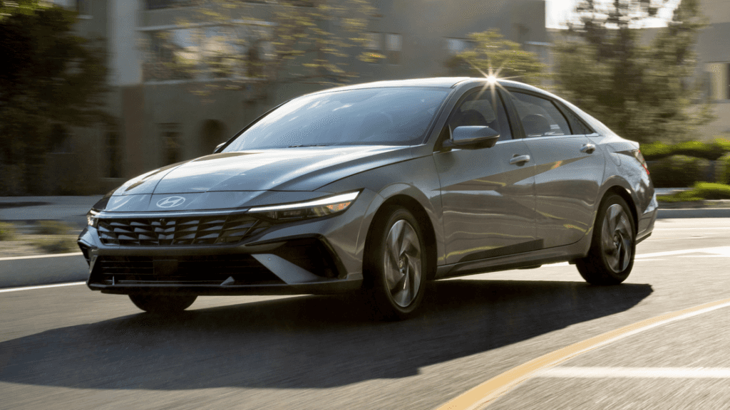 Front-side view of a silver 2025 Hyundai Elantra Hybrid driving on a curved urban road in sunlight.