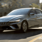 Front-side view of a silver 2025 Hyundai Elantra Hybrid driving on a curved urban road in sunlight.