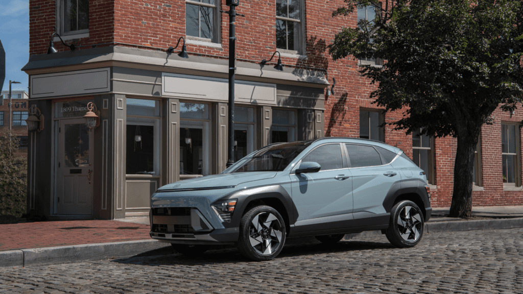 Front-side view of a light blue 2025 Hyundai Kona parked on a cobblestone street beside a brick building.