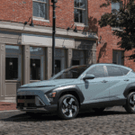 Front-side view of a light blue 2025 Hyundai Kona parked on a cobblestone street beside a brick building.