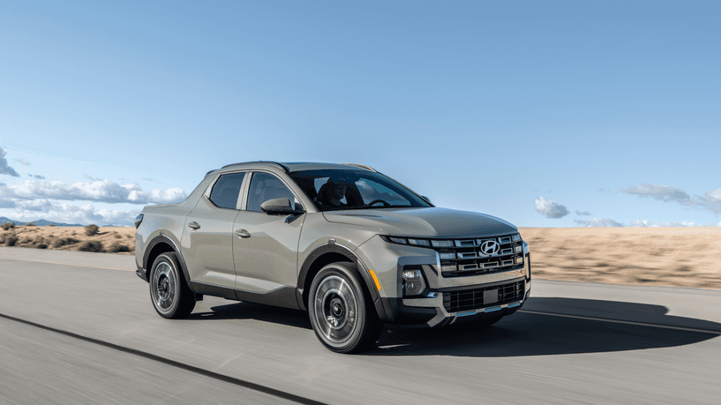 Front-side view of a sage green 2025 Hyundai Santa Cruz driving on an open desert highway under clear skies.