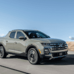 Front-side view of a sage green 2025 Hyundai Santa Cruz driving on an open desert highway under clear skies.