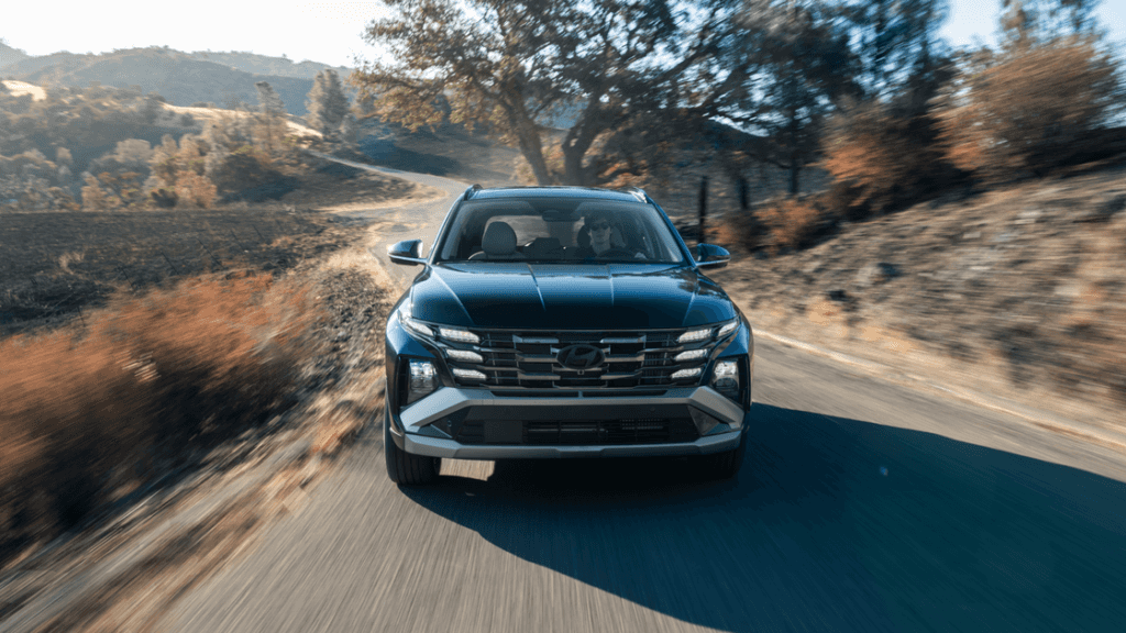 Front view of a black 2025 Hyundai Tucson Plug-in Hybrid driving on a winding rural road with autumn trees.
