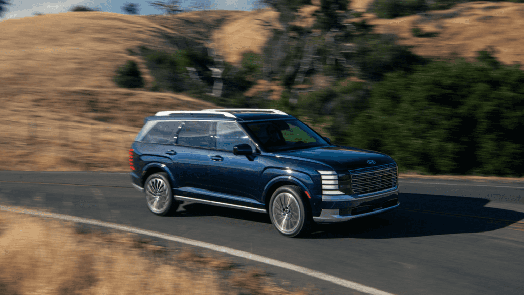 Blue 2026 Hyundai Palisade SUV driving on a winding road with dry hills and greenery in the background, showcasing bold grille and LED headlights.