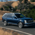 Blue 2026 Hyundai Palisade SUV driving on a winding road with dry hills and greenery in the background, showcasing bold grille and LED headlights.
