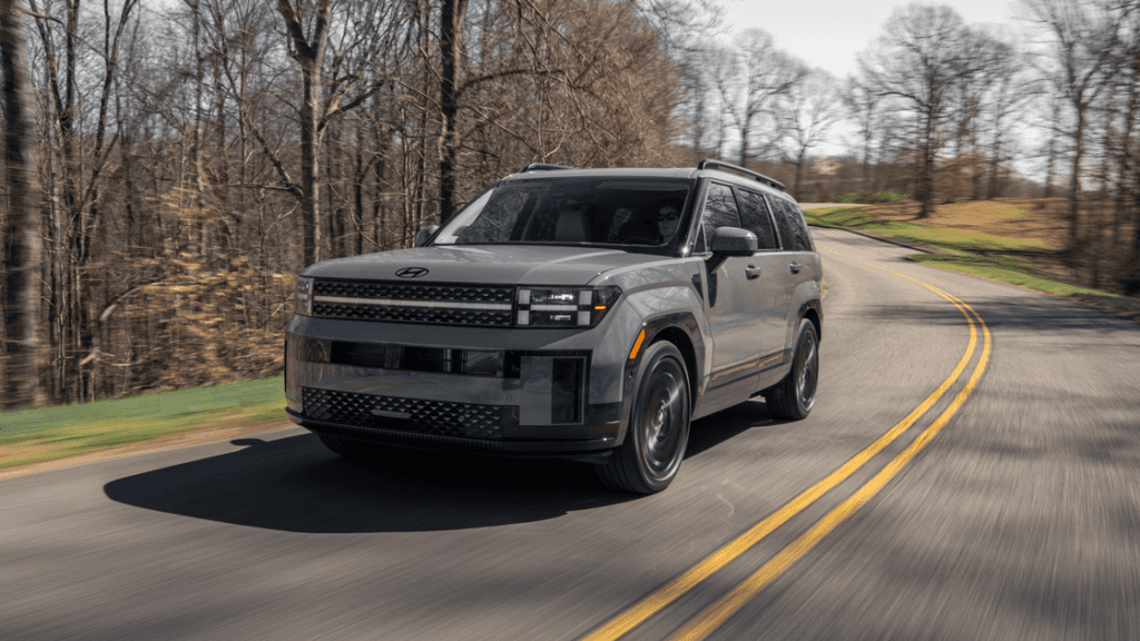 Gray 2026 Hyundai Santa Fe SUV driving on a winding forest road, featuring boxy front fascia, blacked-out grille, and LED headlights.