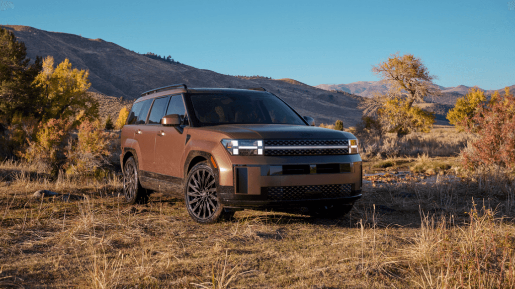 Front view of a bronze 2026 Hyundai Santa Fe Hybrid SUV parked off-road with rugged terrain, showcasing boxy design, LED headlights, and black alloy wheels against a mountain backdrop.