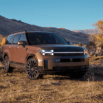 Front view of a bronze 2026 Hyundai Santa Fe Hybrid SUV parked off-road with rugged terrain, showcasing boxy design, LED headlights, and black alloy wheels against a mountain backdrop.