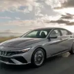 2026 Hyundai Elantra sedan in metallic gray driving on open road with cloudy sky backdrop.