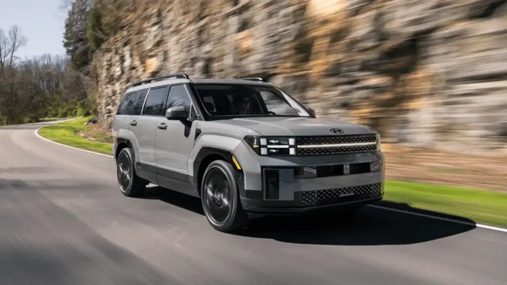 Gray 2025 Hyundai Santa Fe SUV driving through a winding forest road with sunlight reflecting on its body.