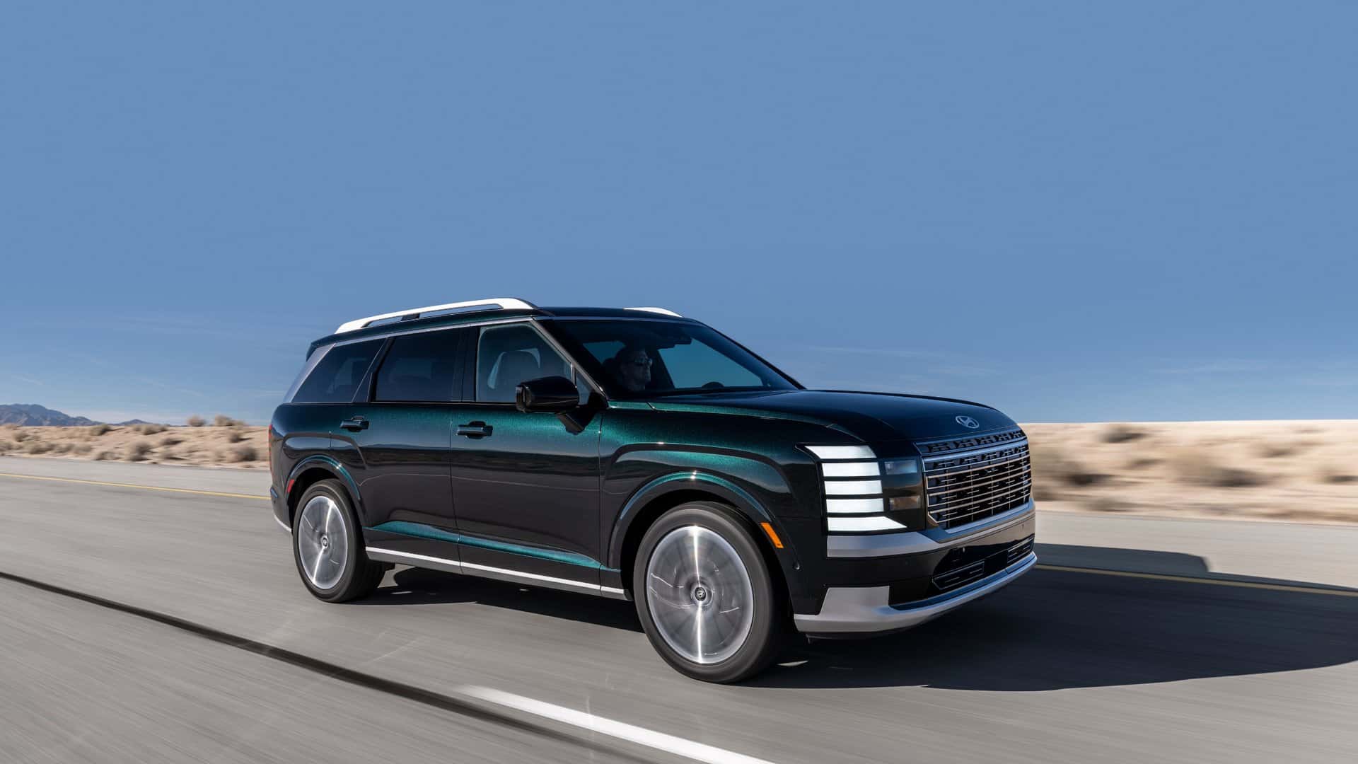 2026 Hyundai Palisade SUV driving on a rural road, showcasing bold grille design, LED lighting, and spacious profile.