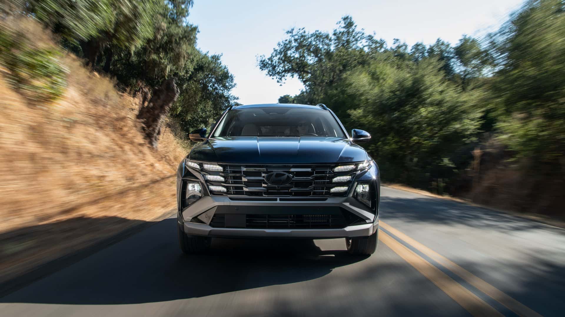 2026 Hyundai Tucson Hybrid front view driving on mountain road with forest scenery.
