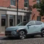 2026 Hyundai Kona green compact SUV parked on cobblestone street near brick building.