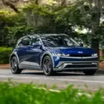 2026 Hyundai IONIQ 5 blue electric SUV driving on tree-lined road with modern pixel lighting.