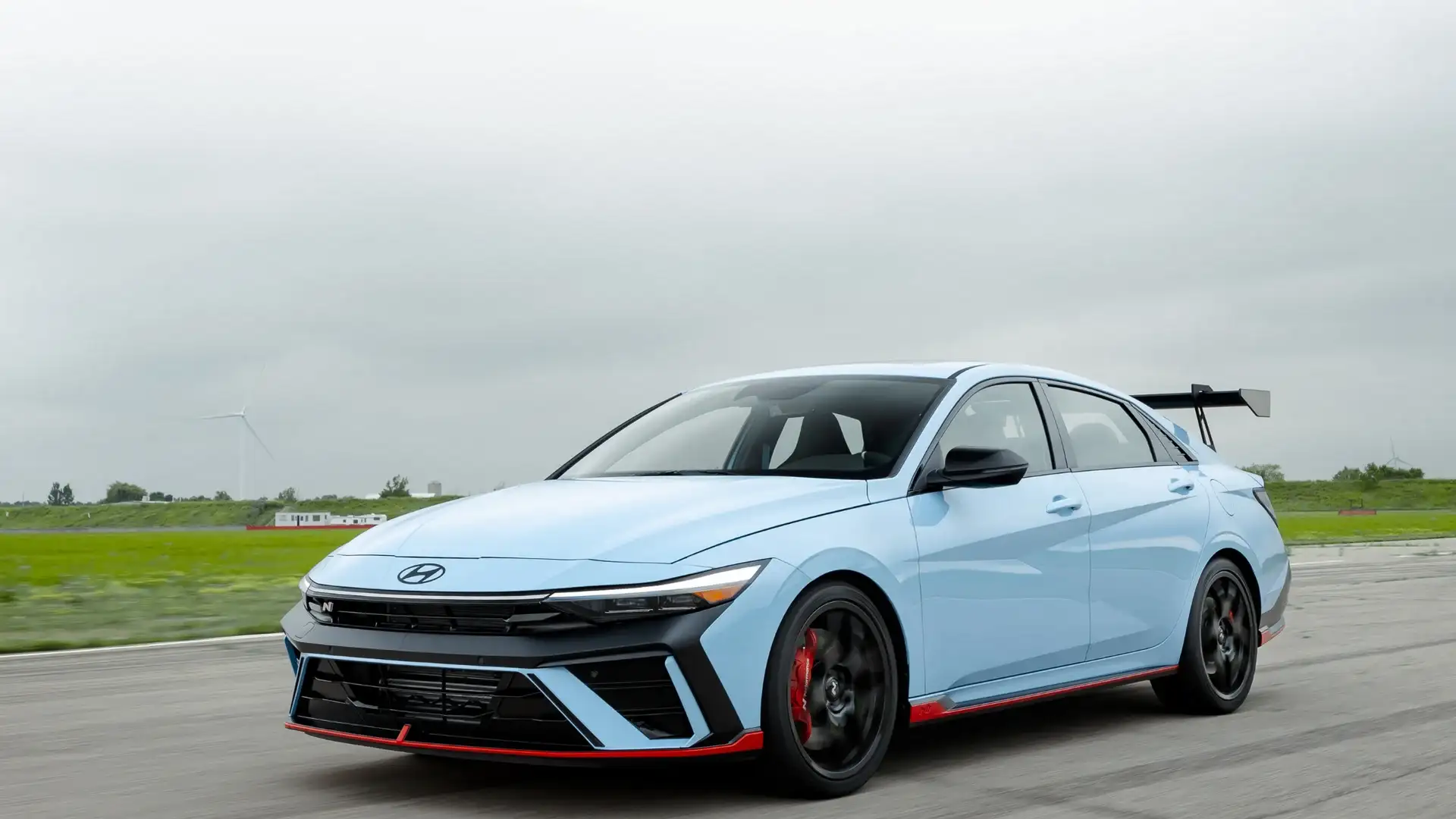 2026 Hyundai Elantra N blue sport sedan driving on track with rear wing and red lower trim.