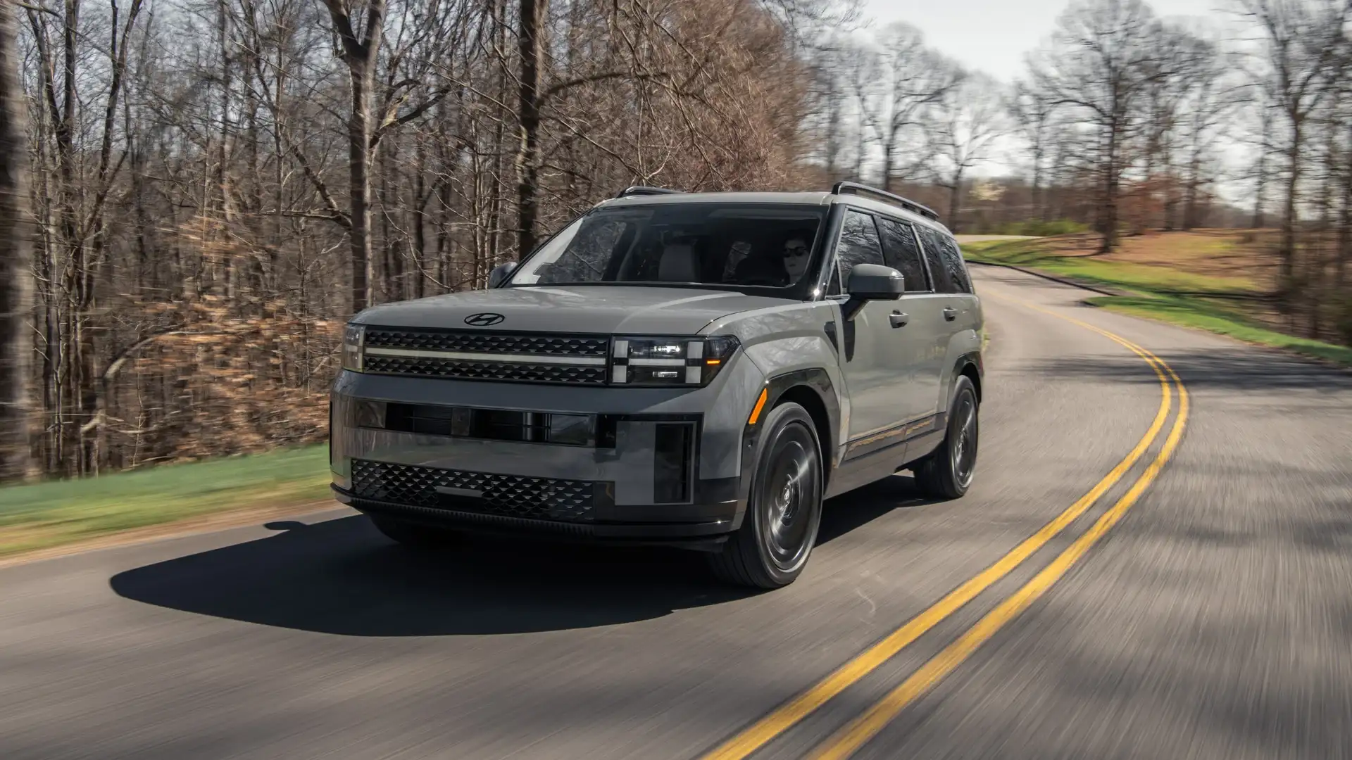 2026 Hyundai Santa Fe gray SUV driving on wooded road with boxy body design and black wheels.