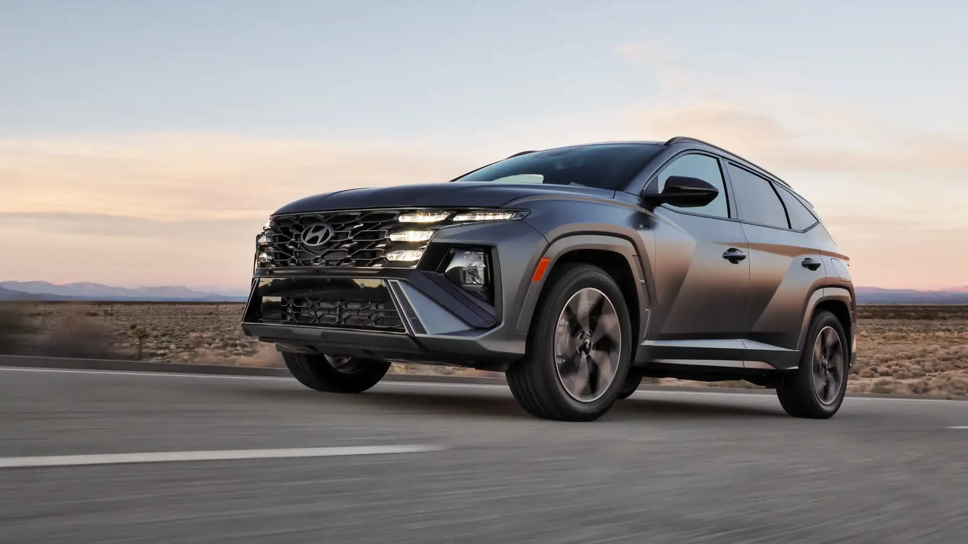 2026 Hyundai Tucson gray SUV driving on desert road at sunset with LED headlights and bold grille.