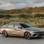 2026 Hyundai Elantra bronze sedan parked on roadside with mountain hills and cloudy sky.