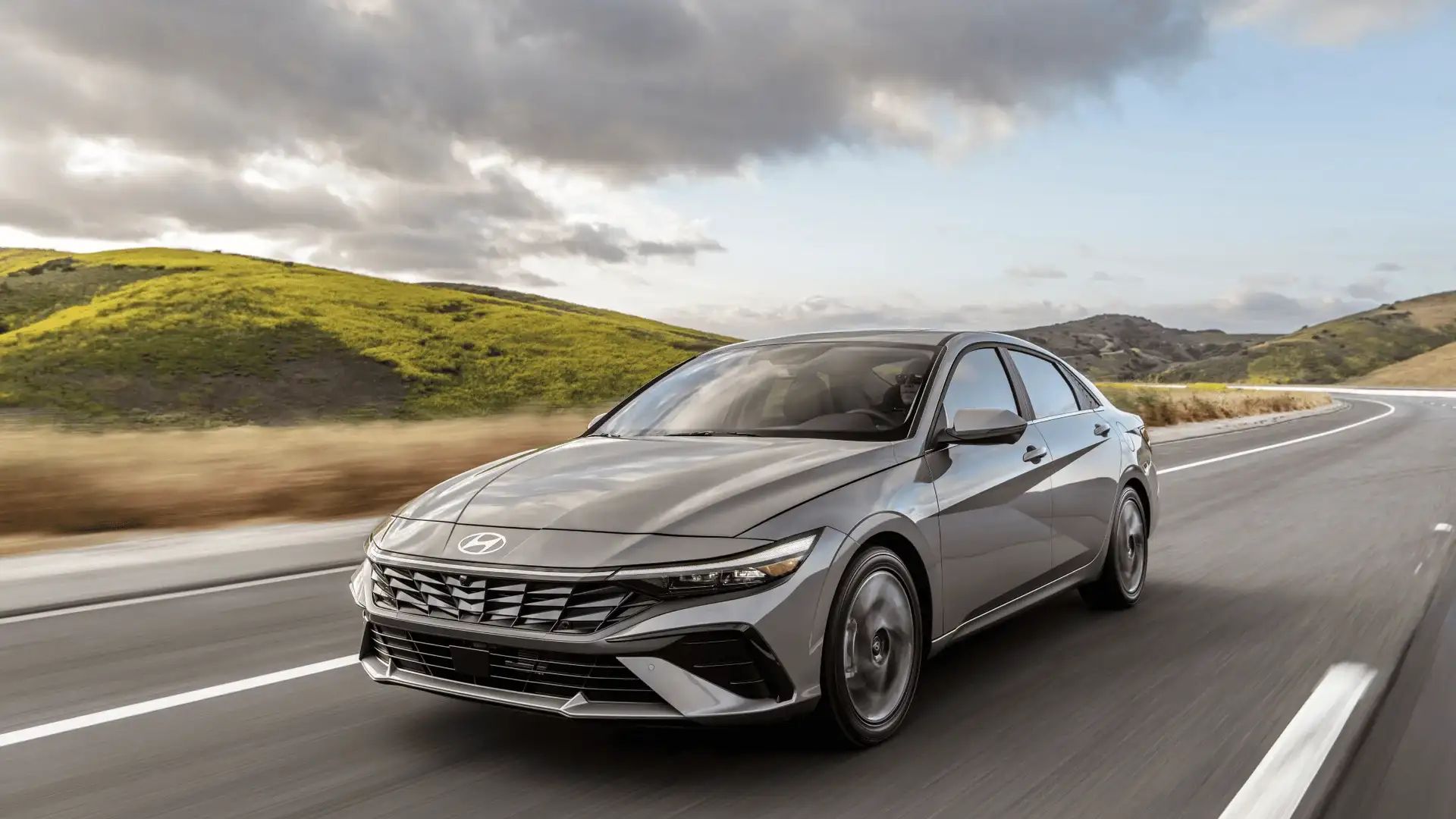 2026 Hyundai Elantra gray sedan driving on open road with hills and cloudy sky.