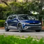 2026 Hyundai IONIQ 5 blue electric SUV driving through tree-lined road with LED headlights.