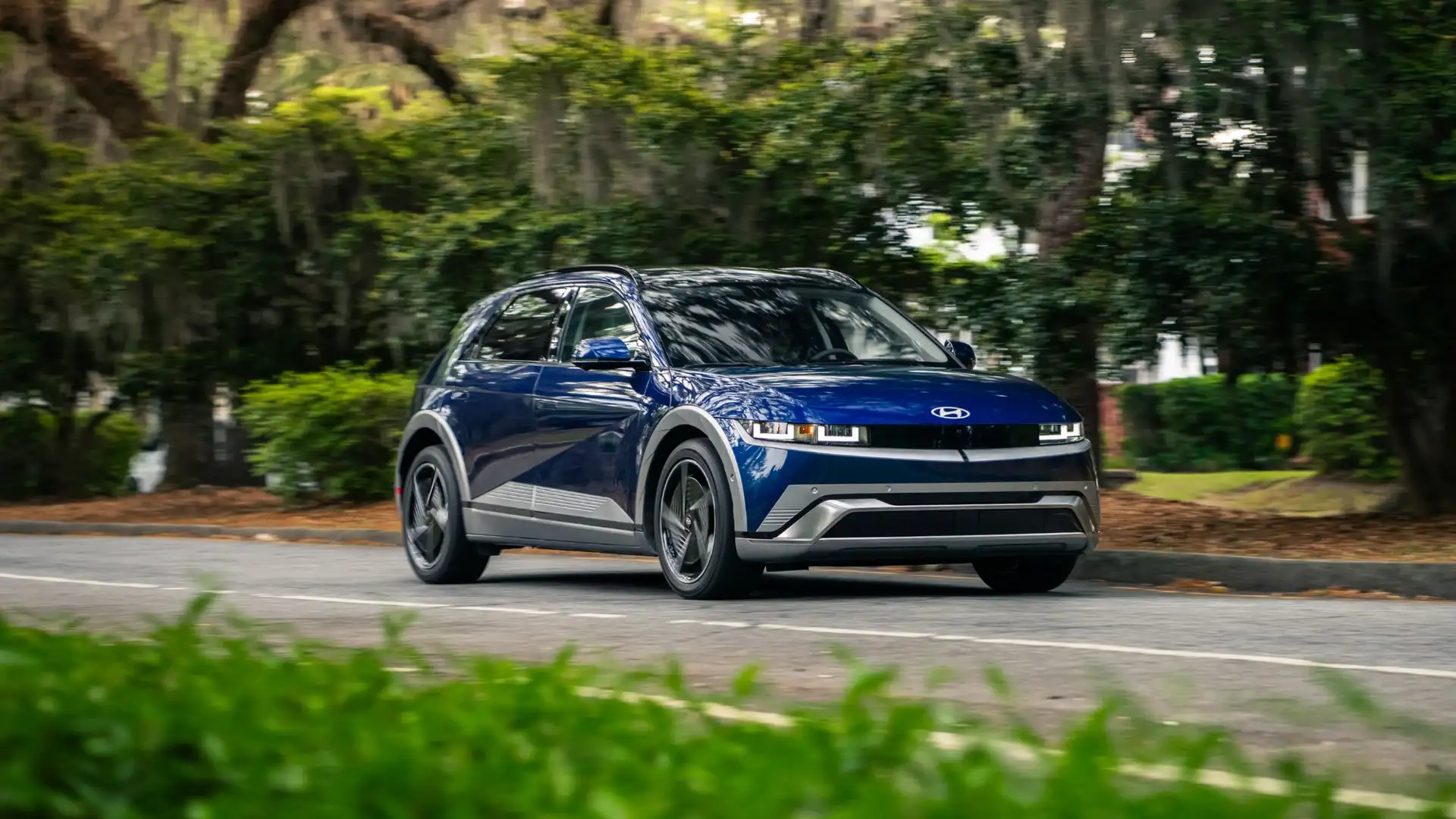 2026 Hyundai IONIQ 5 blue electric SUV driving through tree-lined road with LED headlights.