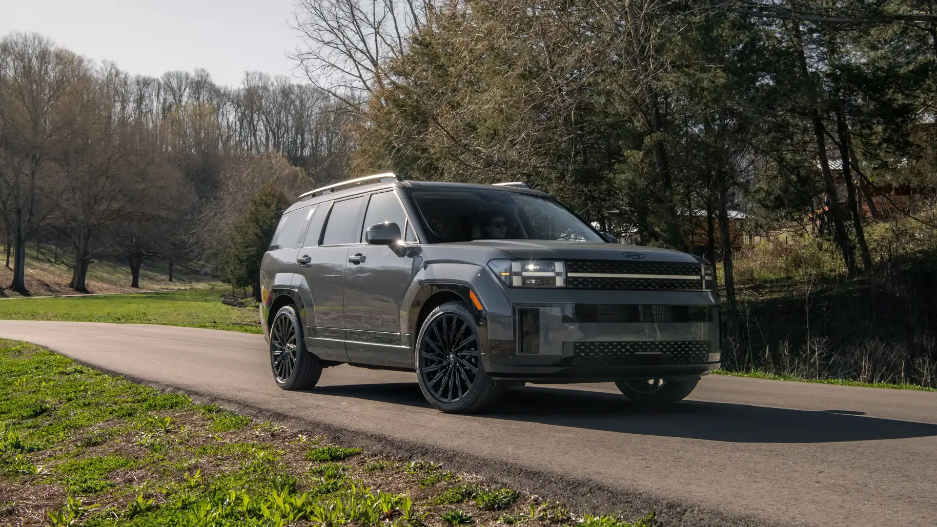 2027 Hyundai Santa Fe gray SUV driving on paved road near trees with front LED lighting.