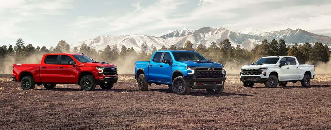 A red, a blue, and a white 2024 Chevy Silverado 1500 are parked on a dirt area.