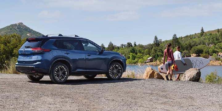 blue 2024 nissan rogue parked on gravel with a man and woman holding a surfboard going into the water. 
