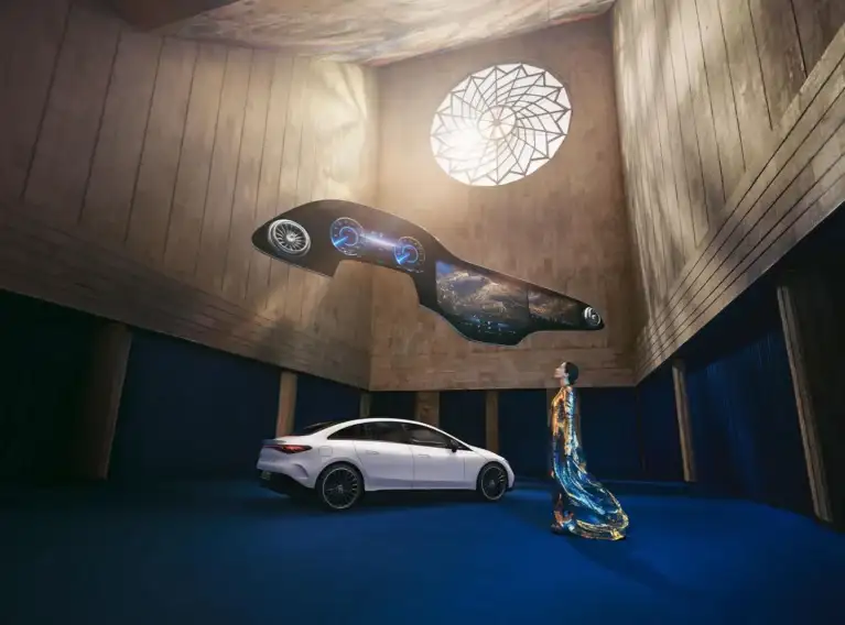 A futuristic Mercedes-Benz Hyperscreen floating above a white Mercedes-Benz electric sedan in an architectural hall with a woman looking up.