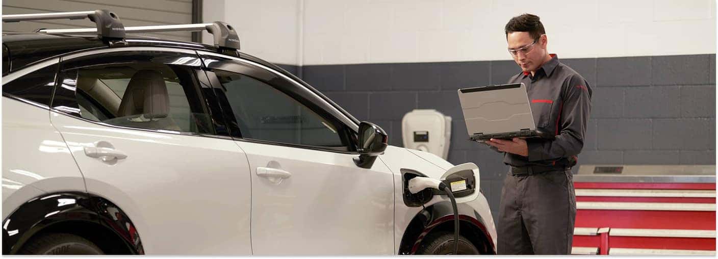A Nissan service technician using a laptop stands next to a white Nissan vehicle connected to a charging cable inside a service bay.