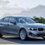 A gray 2026 BMW 3 Series sedan is parked on an asphalt lot, viewed from a front three-quarter angle with a scenic backdrop of misty mountains and trees under a cloudy sky.