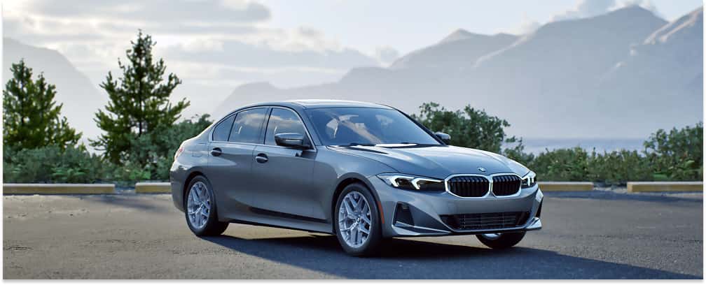 A gray 2026 BMW 3 Series sedan is parked on an asphalt lot, viewed from a front three-quarter angle with a scenic backdrop of misty mountains and trees under a cloudy sky.