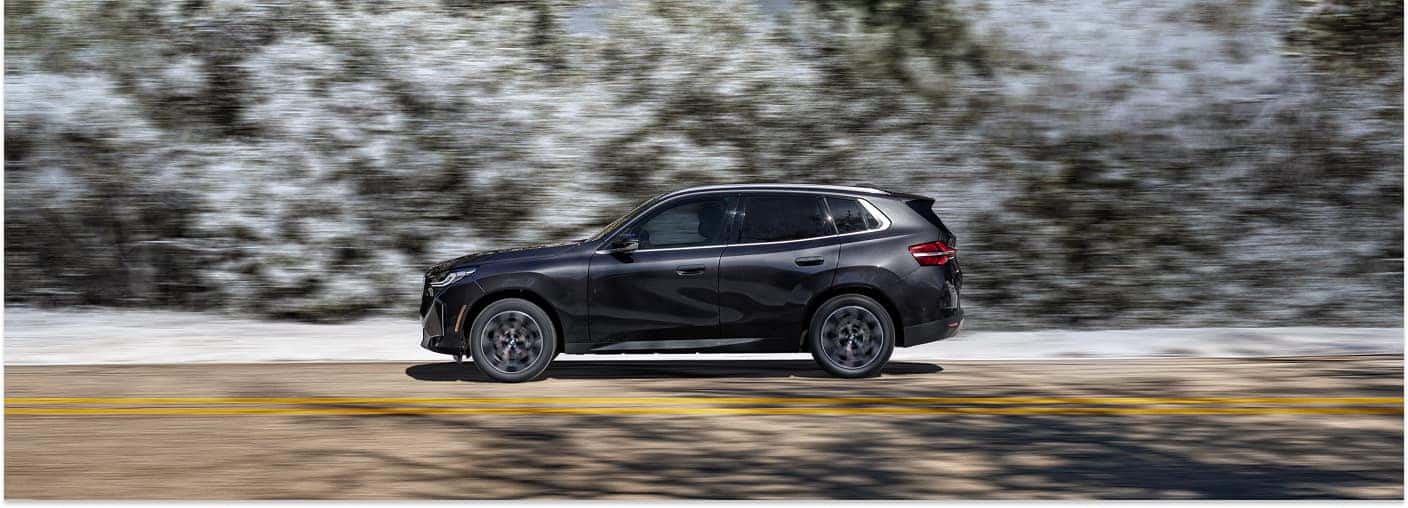 Side profile of a dark metallic 2026 BMW X3 driving on a paved road.