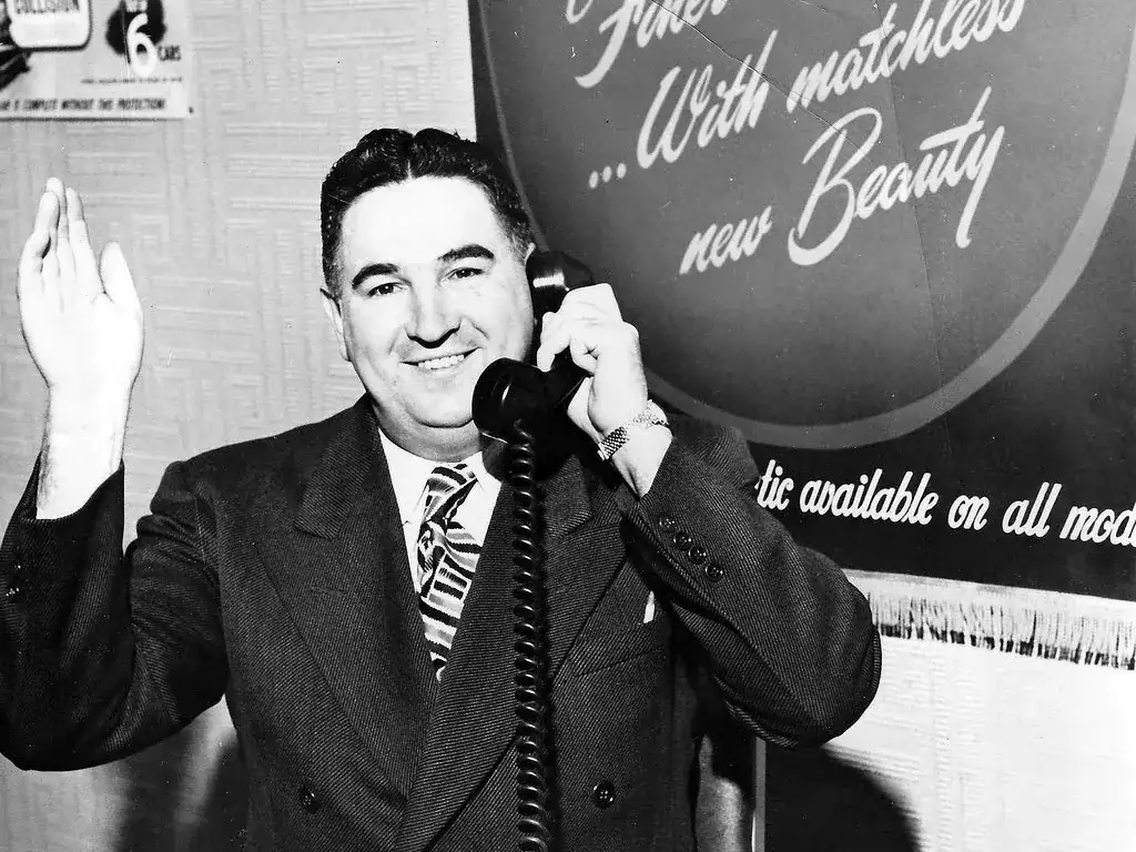 Bud Clary, smiling, on the telephone at his desk with one hand raised, in a black and white photo
