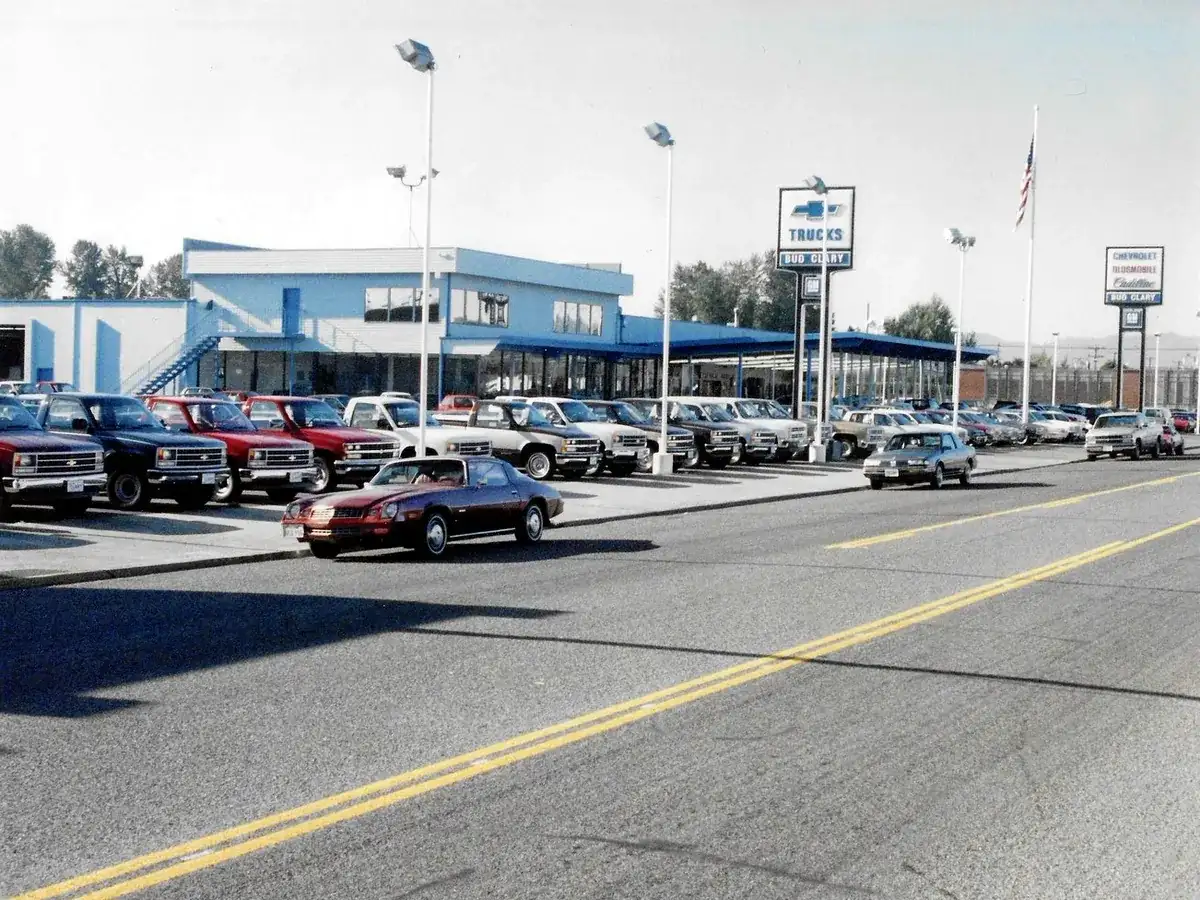 A row of new trucks and cars along the street in front of a blue and white Bud Clary store with Chevrolet, Oldsmobile and Cadillac signs, in a faded color photo