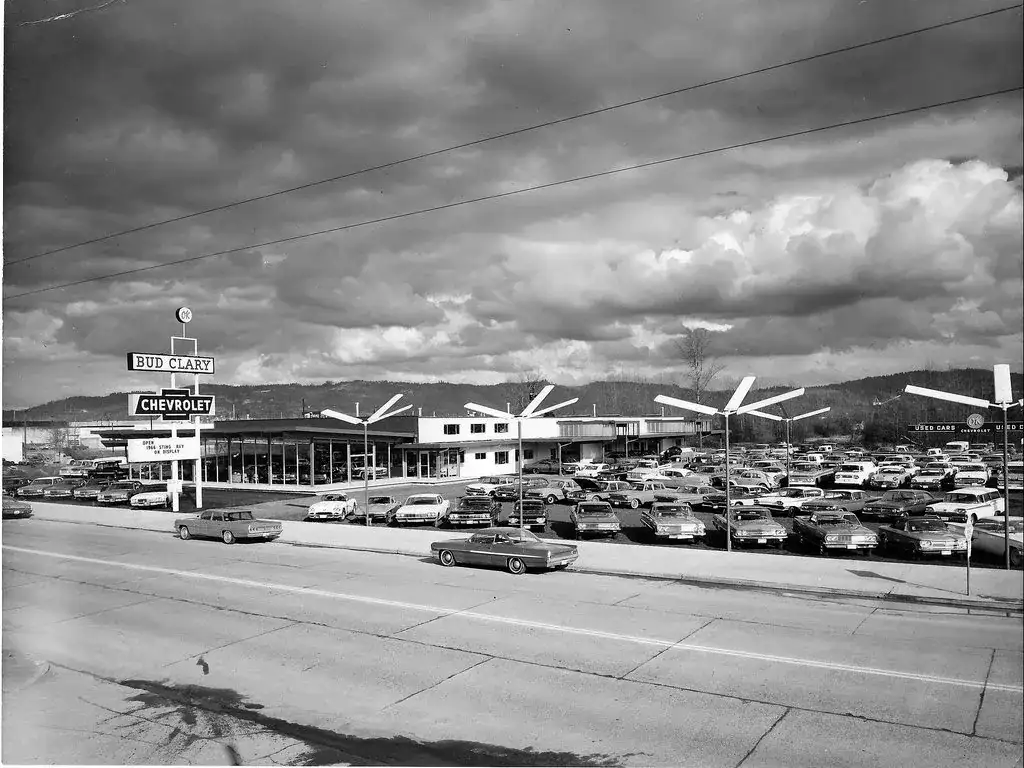 Bud Clary Chevrolet's showroom and a lot full of 1960s cars under a cloudy sky, with a reader board announcing a 1966 Sting Ray on display, in a black and white photo