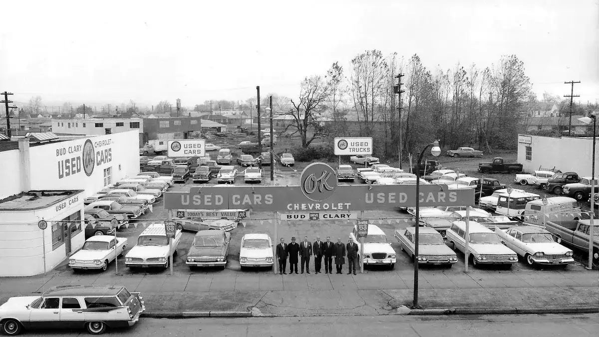Rows of cars under an OK Used Cars sign at Bud Clary Chevrolet, with the sales staff lined up in dark coats, in a black and white photo
