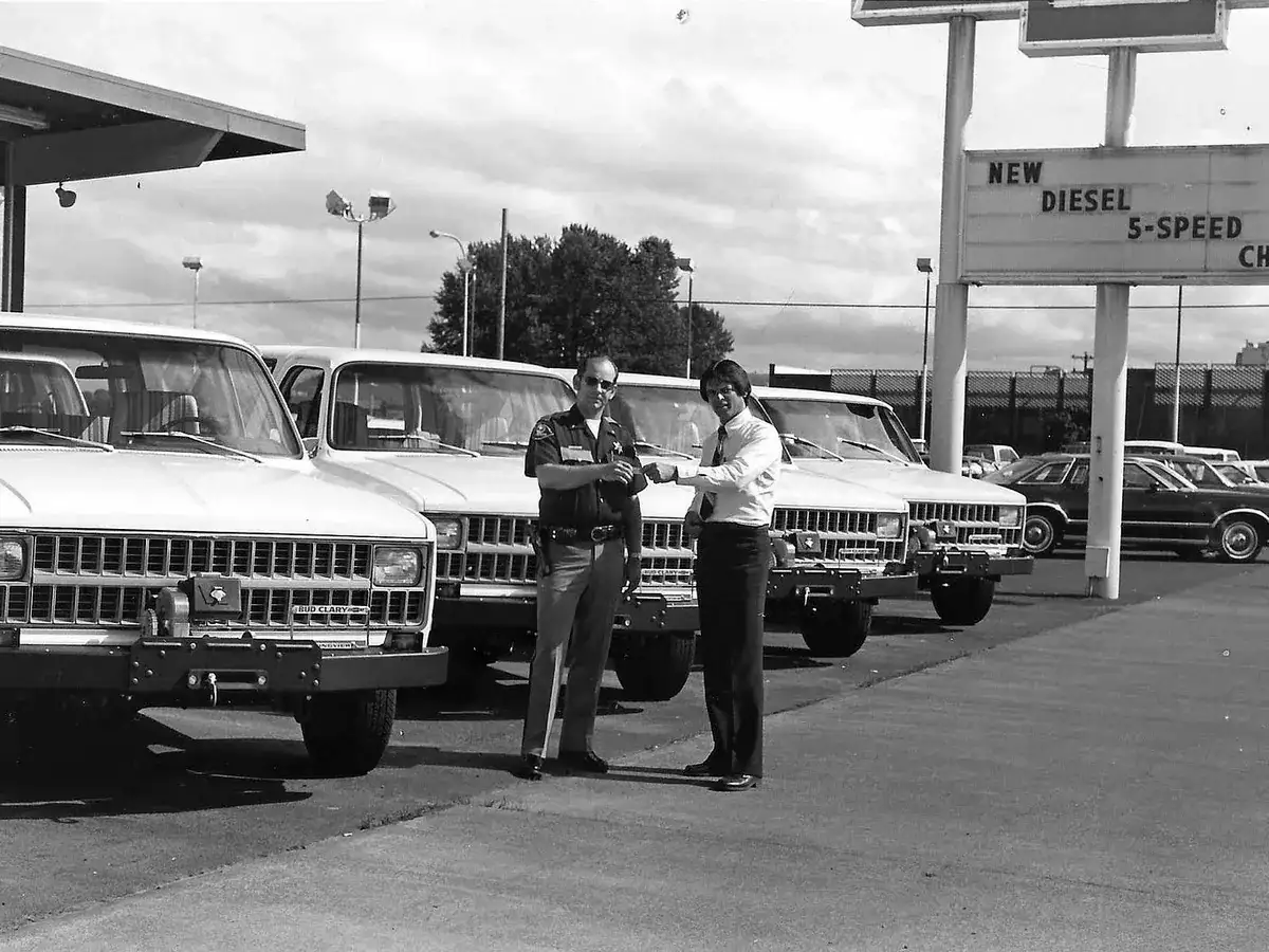 A sheriff's deputy and a man in a white shirt beside a row of new Chevrolet Blazers fitted with winches, in a black and white photo