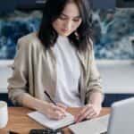 Young woman writing on a notepad at her desk with a laptop and calculator.