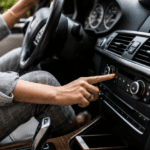 Close-up of a person inside a modern car adjusting the dashboard audio or climate controls. One hand reaches toward the center console while the steering wheel, air vents, digital display, gear selector, and part of a smartphone in a storage compartment are visible. The image is shot with shallow depth of field, emphasizing the hand and control panel.