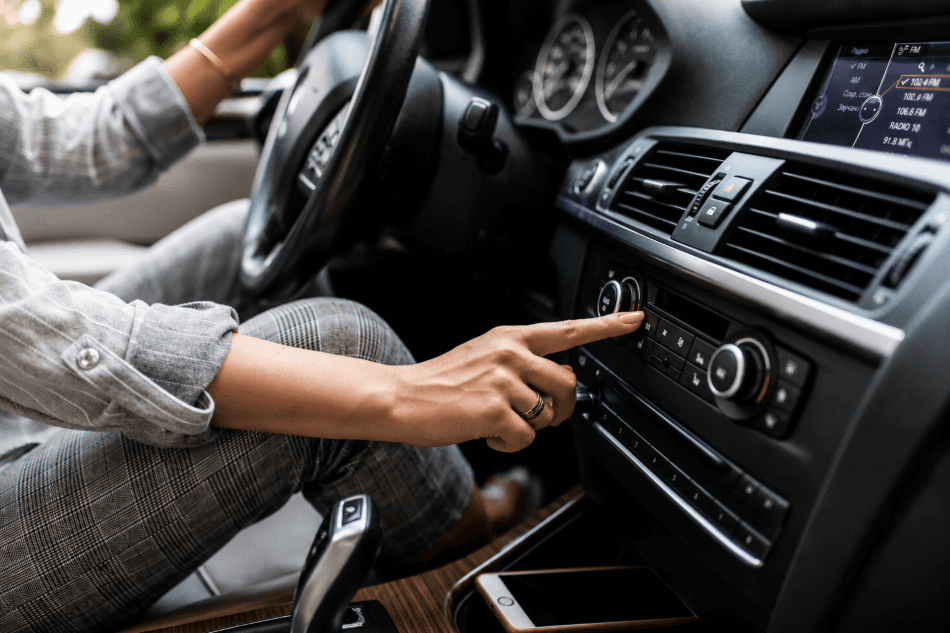 Close-up of a person inside a modern car adjusting the dashboard audio or climate controls. One hand reaches toward the center console while the steering wheel, air vents, digital display, gear selector, and part of a smartphone in a storage compartment are visible. The image is shot with shallow depth of field, emphasizing the hand and control panel.