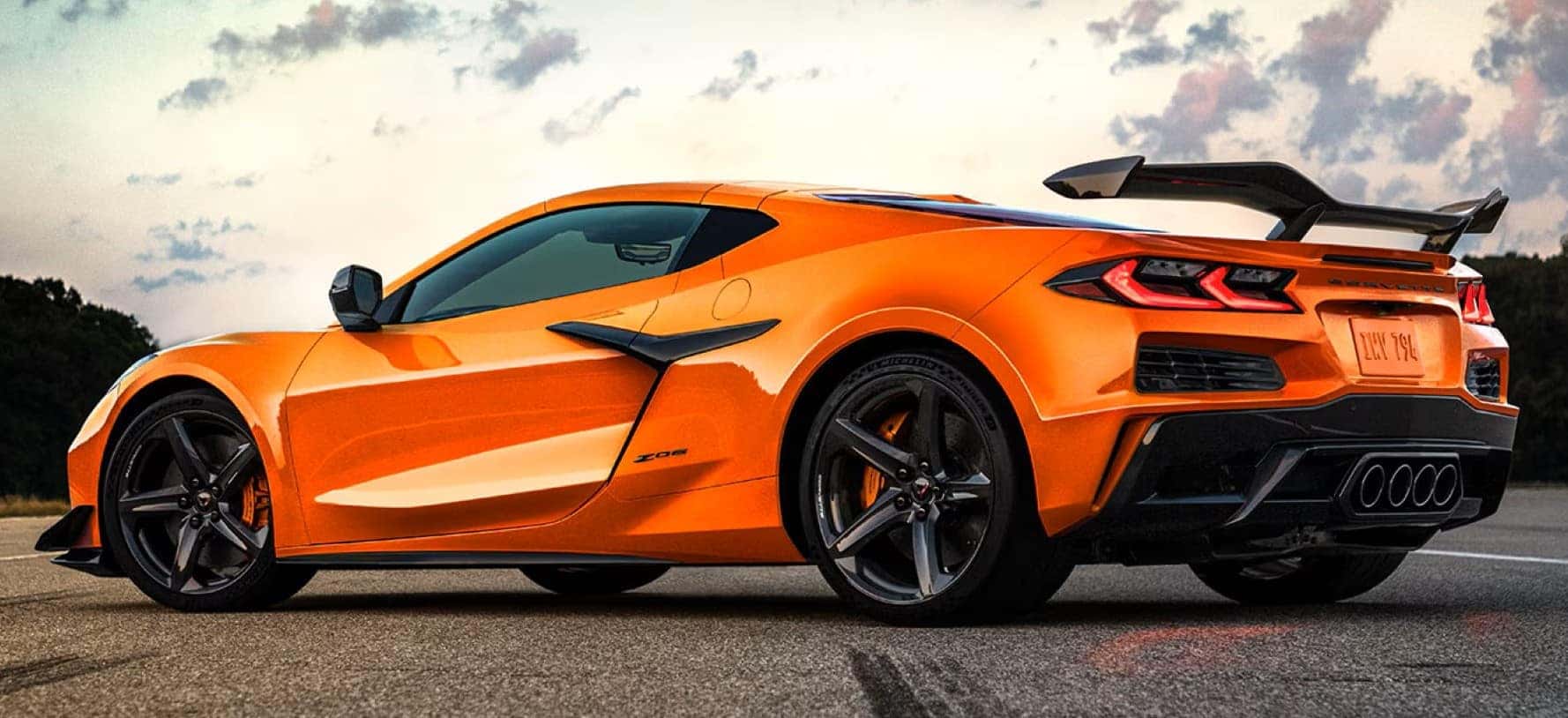 Rear-side view of an orange 2025 Chevrolet Corvette Z06 parked on pavement, featuring a rear spoiler, quad exhaust tips, and black alloy wheels.