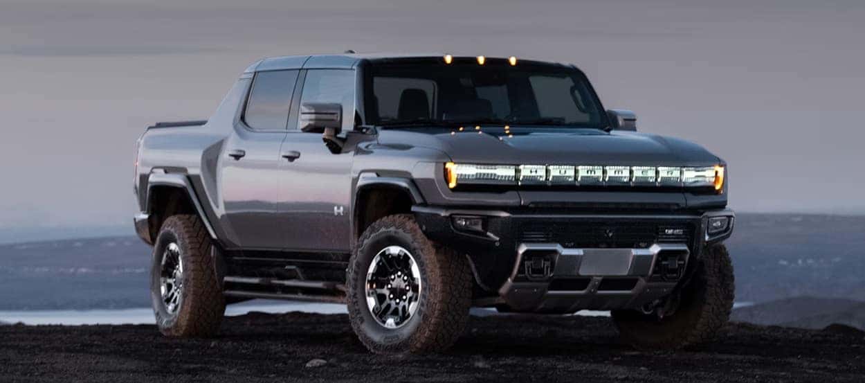 Dark gray 2025 GMC HUMMER EV Pickup parked on rocky terrain at dusk, with headlights illuminated and a scenic landscape in the background.