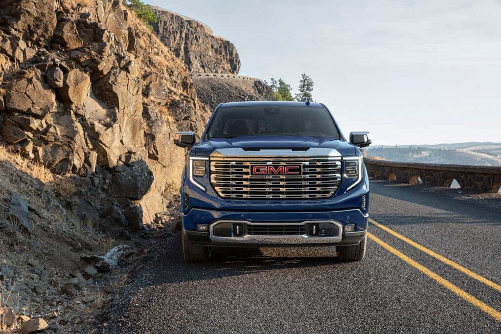 Front view of a blue 2025 GMC Sierra 1500 parked on a mountain road with rocky cliffs and scenic views in the background.