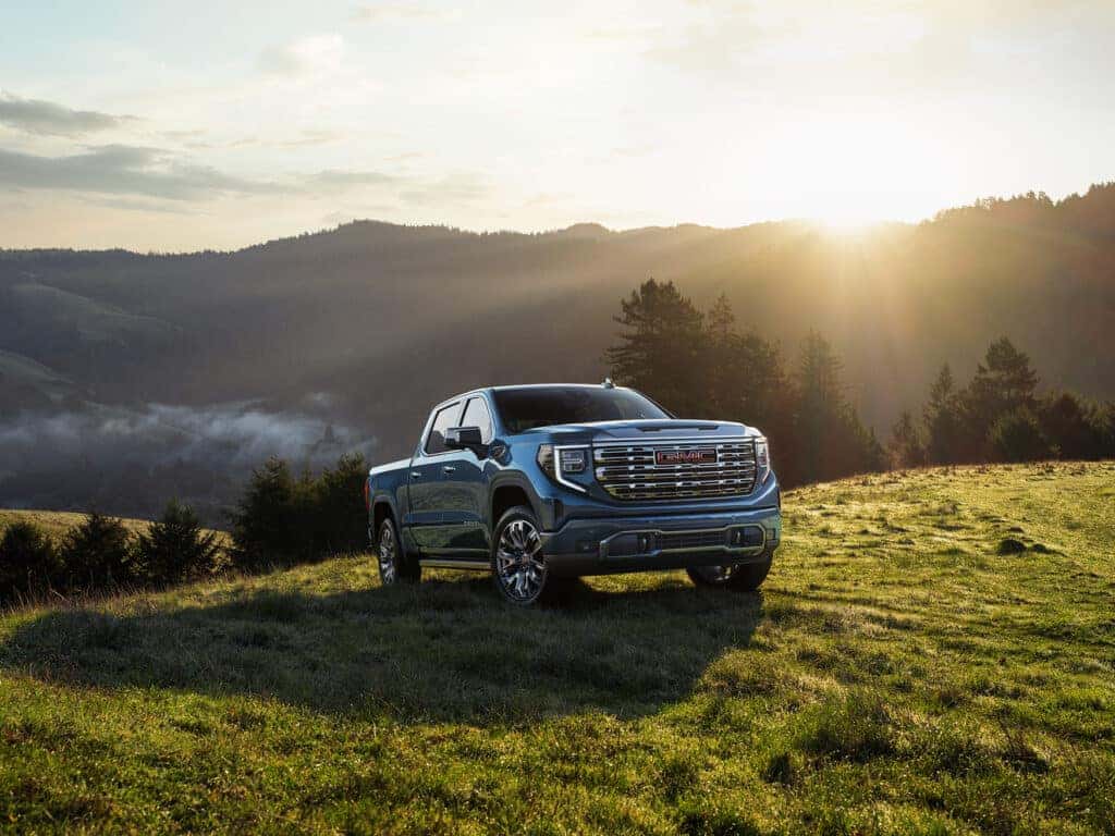 2025 GMC Sierra 1500 parked on a grassy hill at sunrise, with a scenic view of foggy mountains and forest in the background.