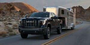 2025 GMC Sierra 2500 in black towing a large silver trailer through a desert mountain road, showcasing heavy-duty capability and bold styling.
