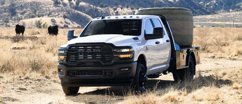 Front three-quarter view of a white RAM Chassis Cab flatbed truck loaded with a hay bale in a field.